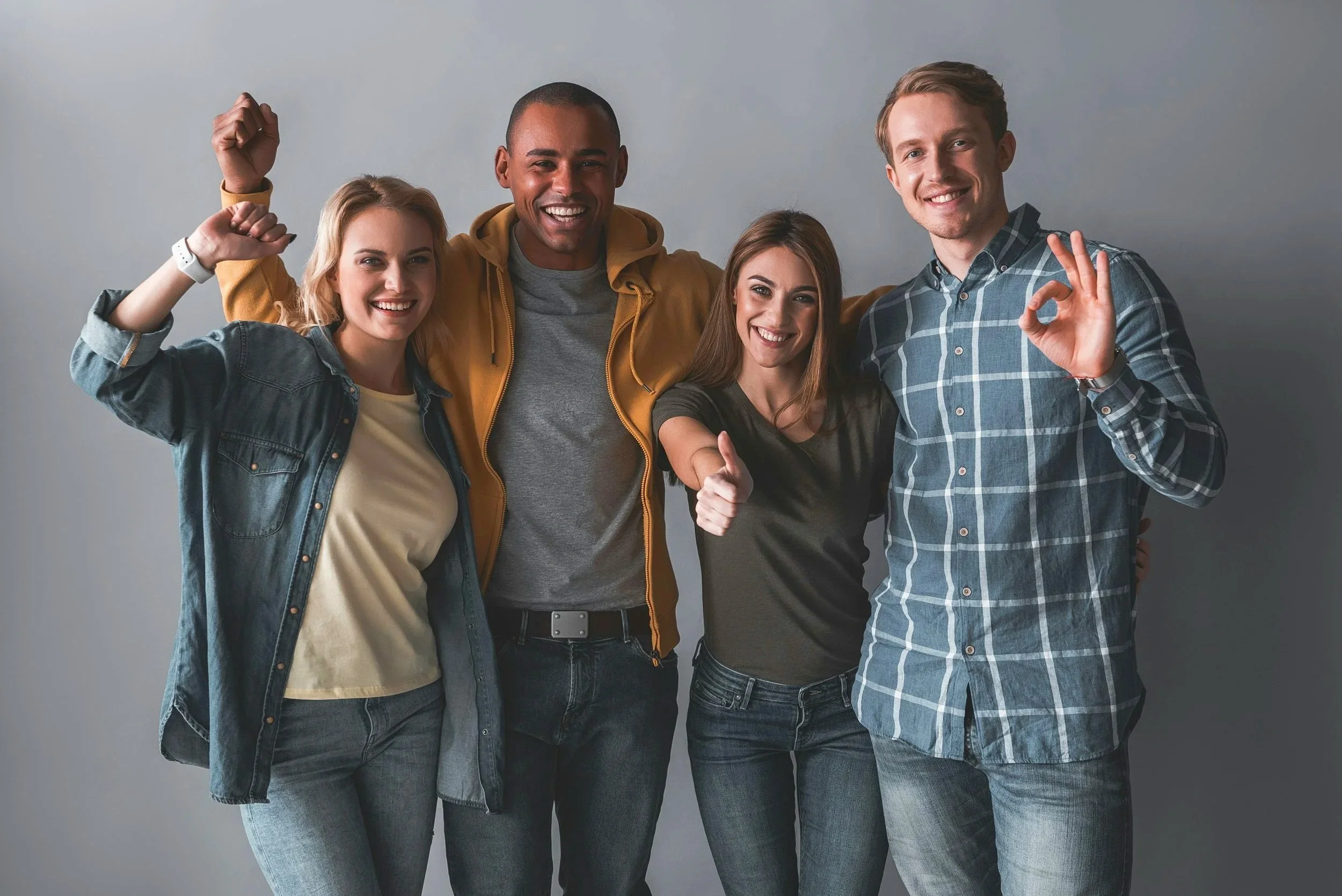 Group of five diverse young adults smiling and posing together, some making gestures like a thumbs-up, arm raised, and an 'OK' sign against a plain gray background.