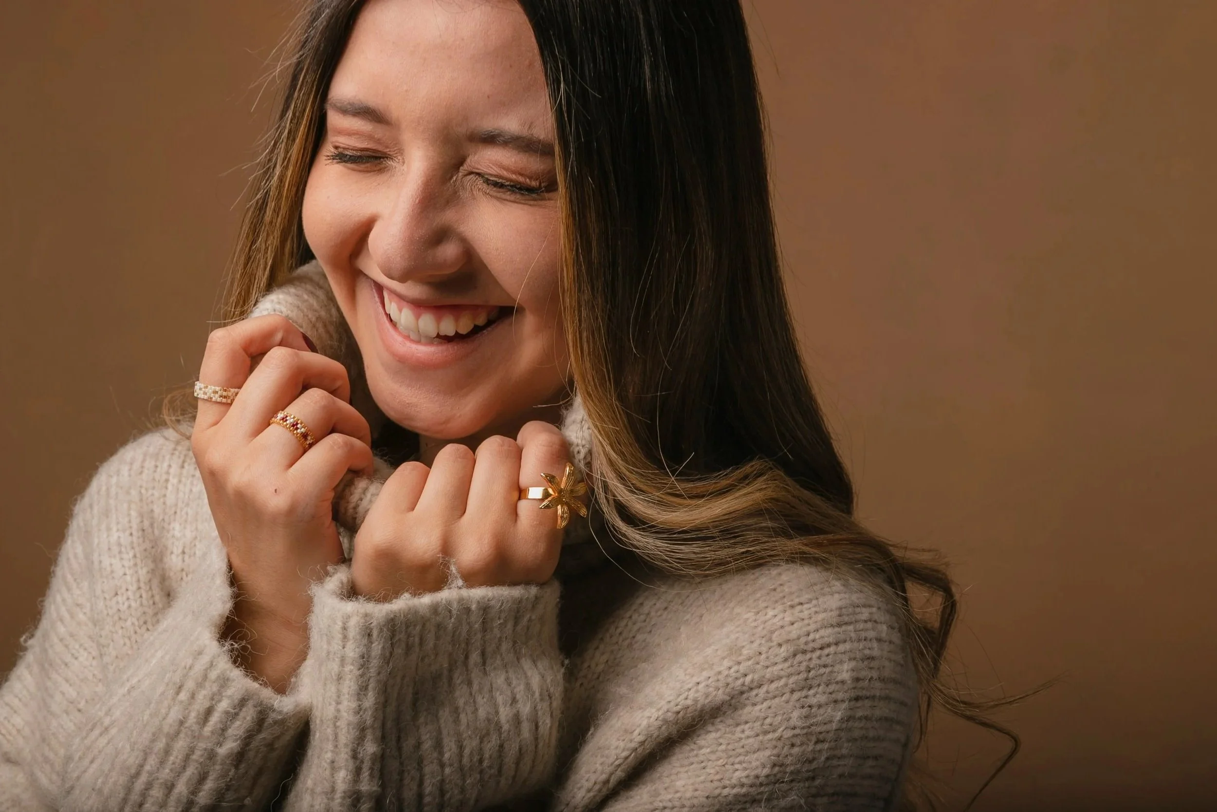 A woman with long wavy hair, smiling with eyes closed, wearing multiple rings and a cozy light-colored sweater, standing against a brown background.