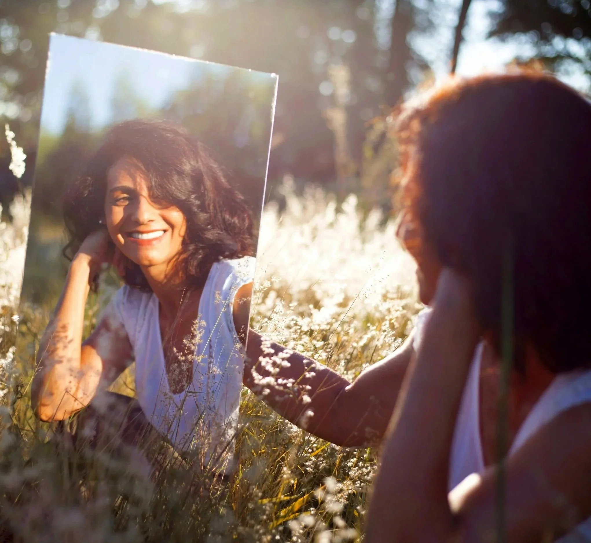 A woman smiling and holding a mirror in a field during sunlight with trees in the background.