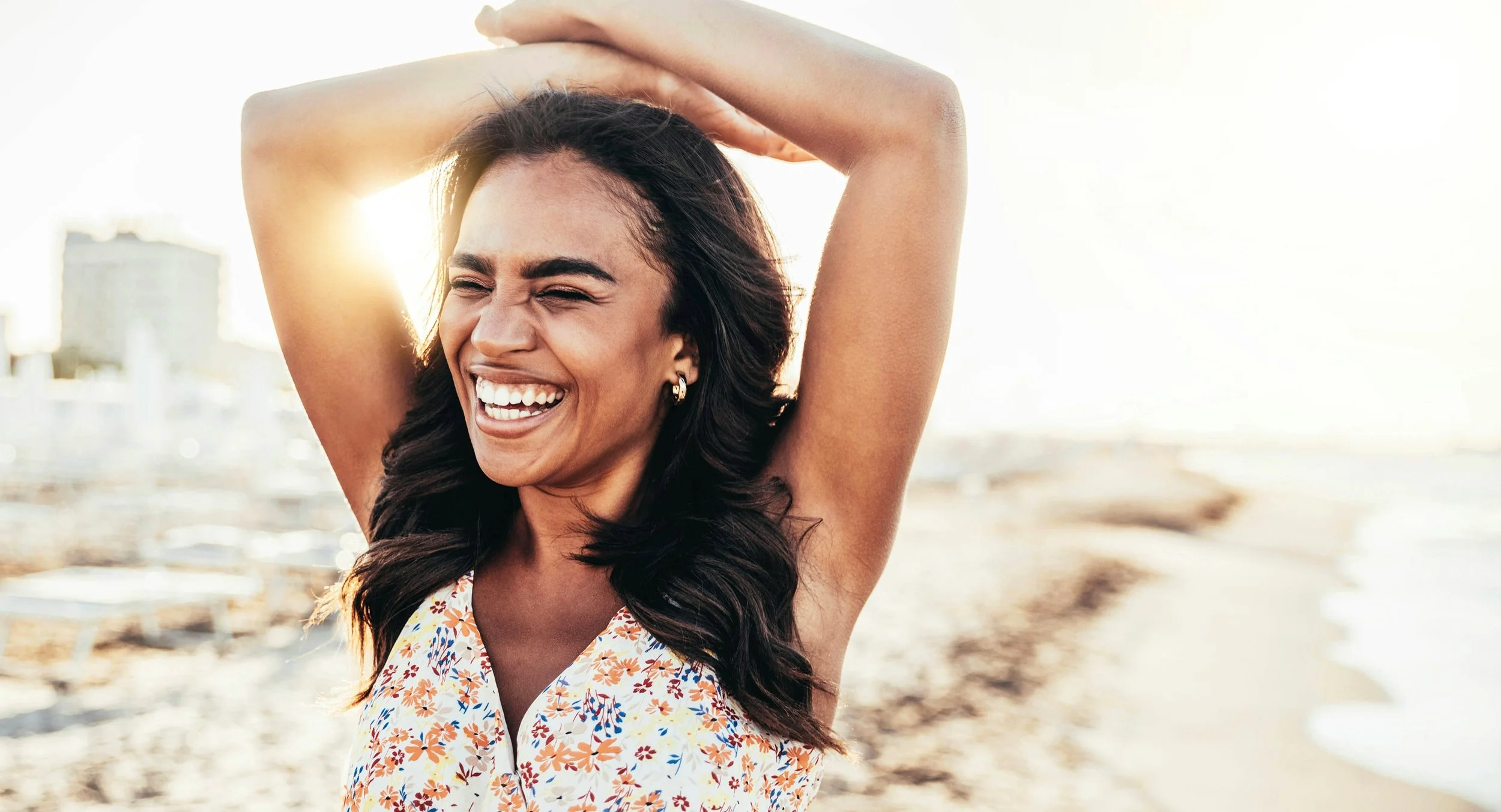 A woman smiling joyfully at the beach during sunset with her arms raised above her head.