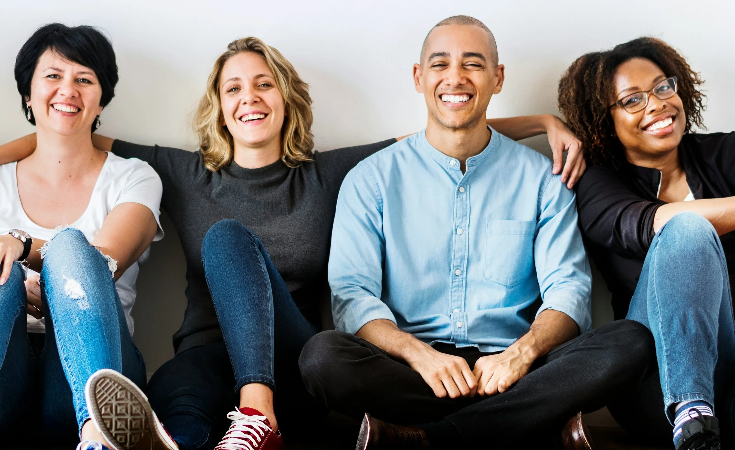 A group of four people sitting close together, smiling, with their arms around each other, against a plain white background.