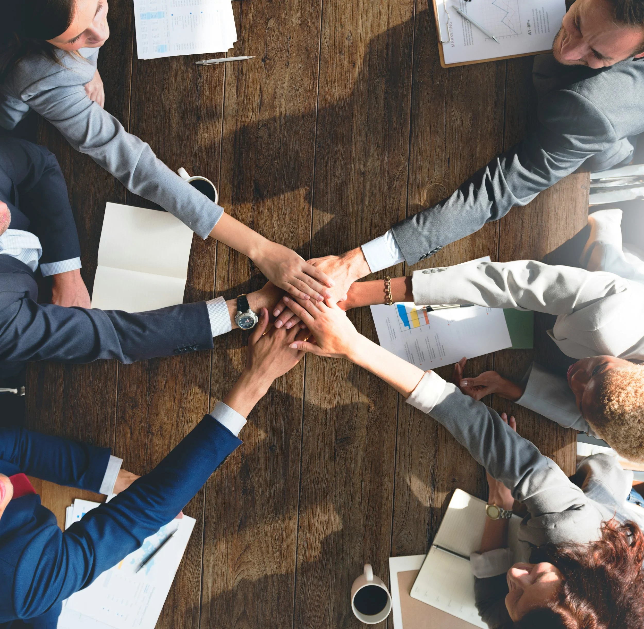 A group of diverse professionals sitting around a wooden table with documents, charts, pens, and coffee cups, engaging in a collaborative meeting.