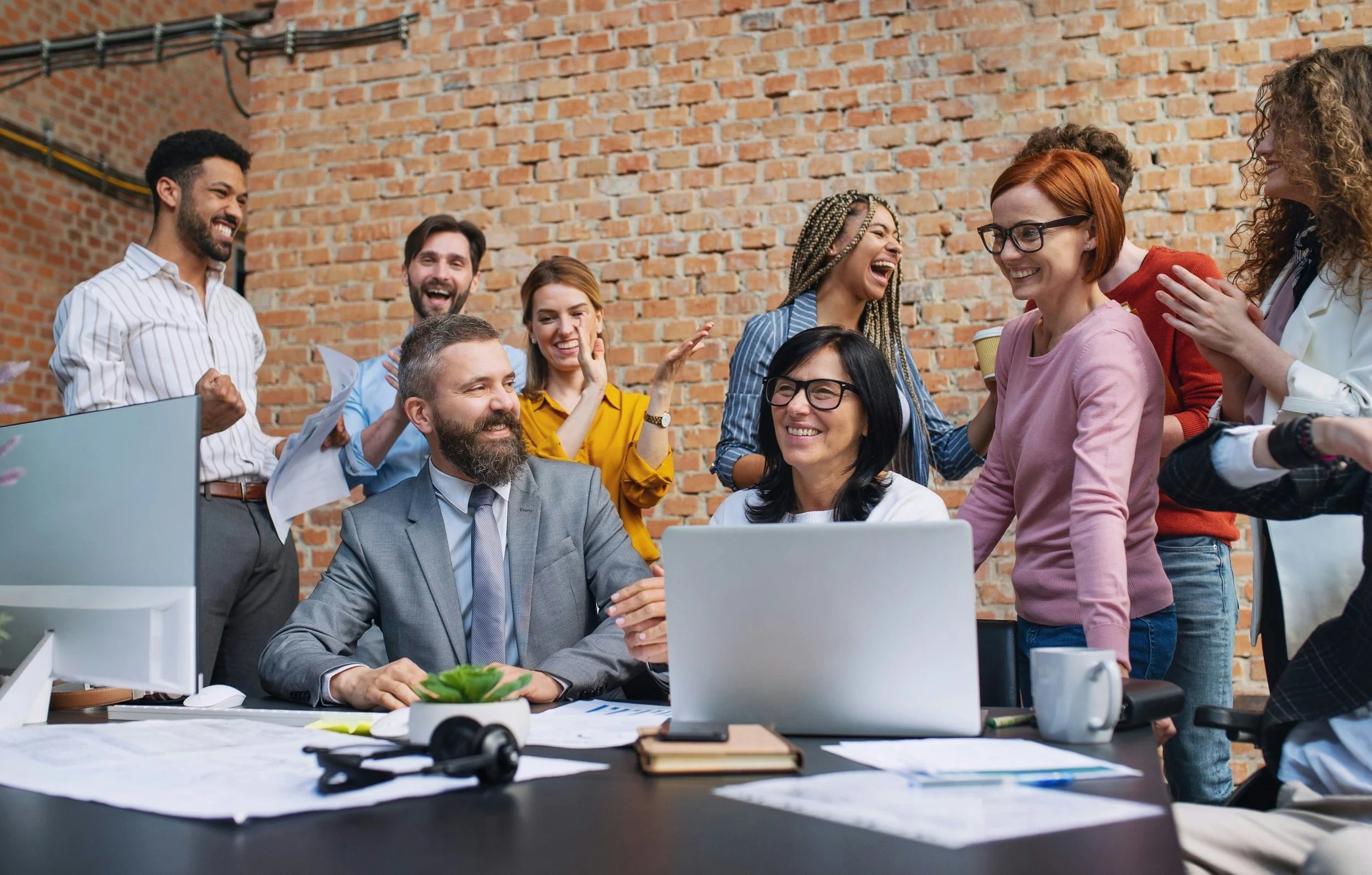 A diverse group of coworkers are gathered around a desk in an office with an exposed brick wall. They are laughing and smiling, enjoying a moment of camaraderie. There are various papers, books, and a laptop on the desk.