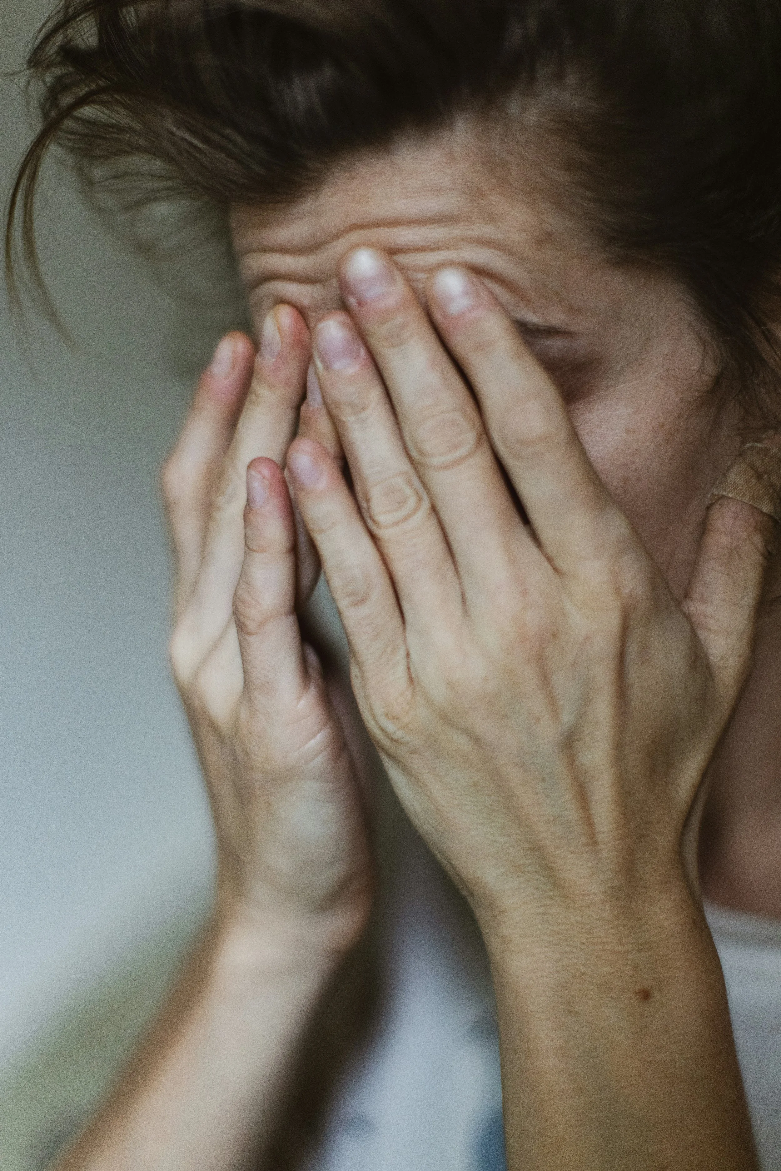 A person with brown hair touching their face with their hands, covering one eye, with a neutral or distressed expression.