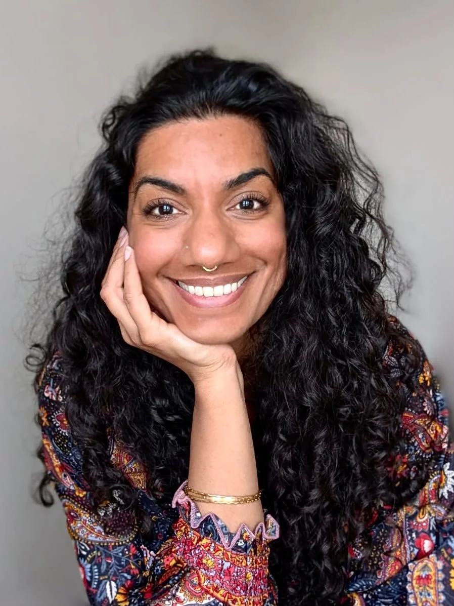 A woman with long, curly black hair smiling and resting her chin on her hand. She is wearing a colorful patterned blouse and gold bracelets, against a neutral background.