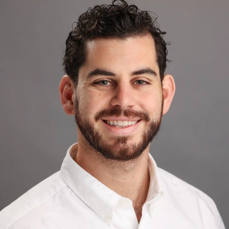 A young man with curly dark hair, a beard, and a friendly smile, wearing a white button-up shirt, posing against a gray background.