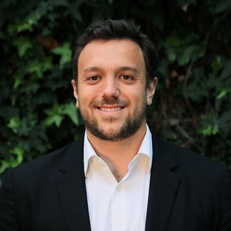 A man with short dark hair and a beard, dressed in a white collared shirt and black blazer, smiling in front of green leafy background.
