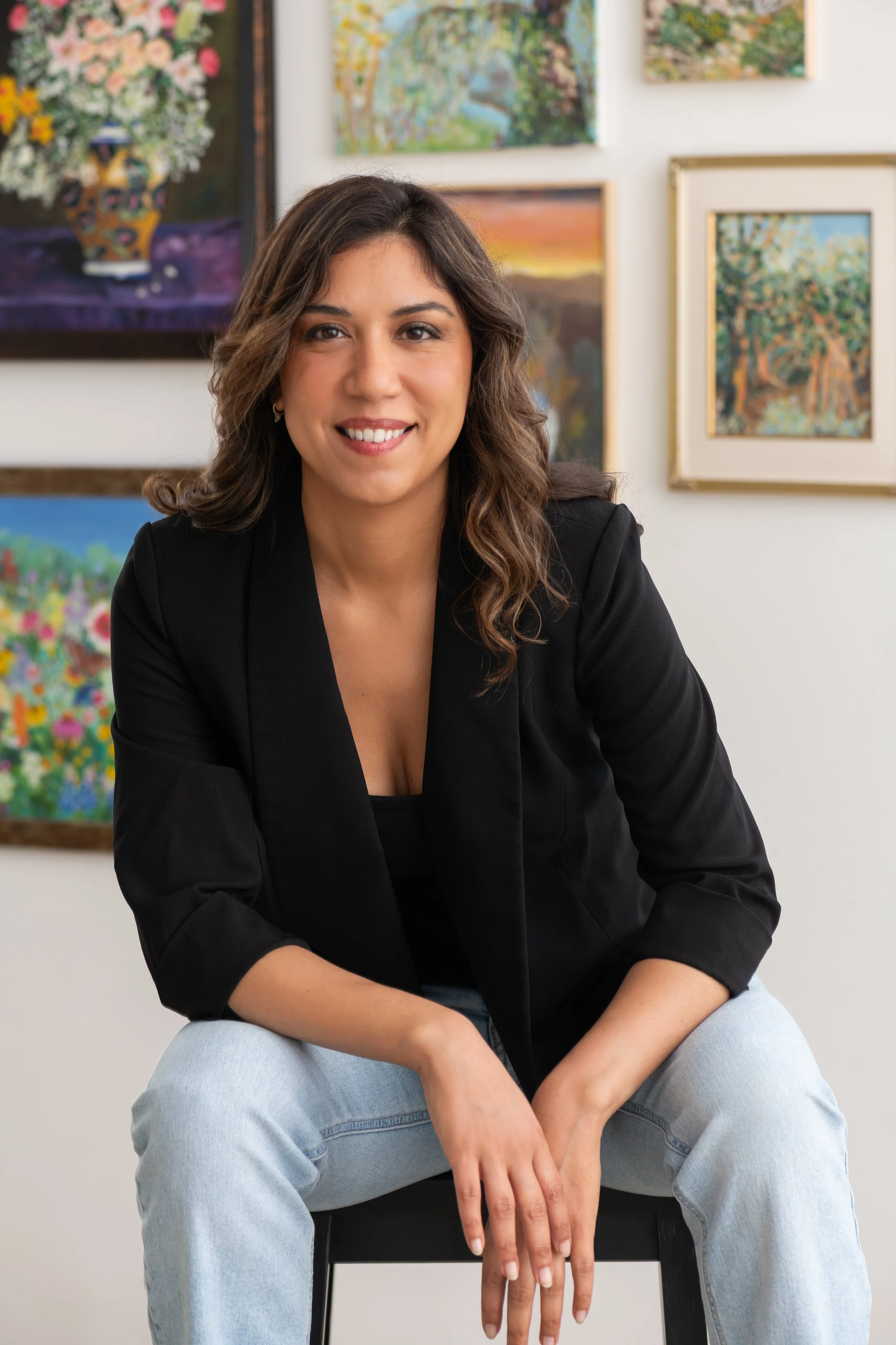 A young woman with wavy brown hair smiling, wearing a black blazer and light blue jeans, sitting on a black chair in an art gallery with colorful paintings on the wall behind her.