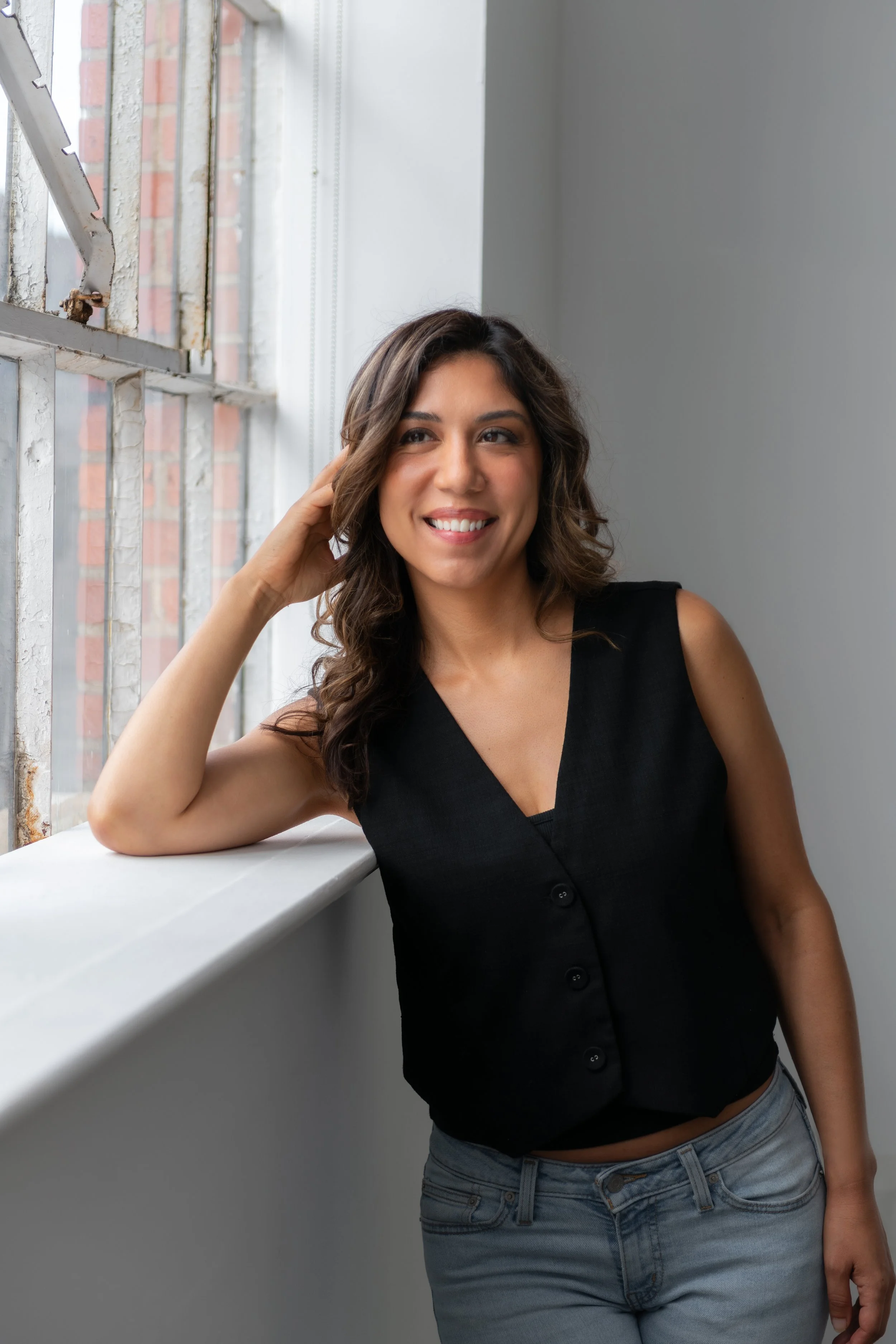 A woman with wavy brown hair wearing a sleeveless black top leaning on a white windowsill, smiling, and looking out the window.