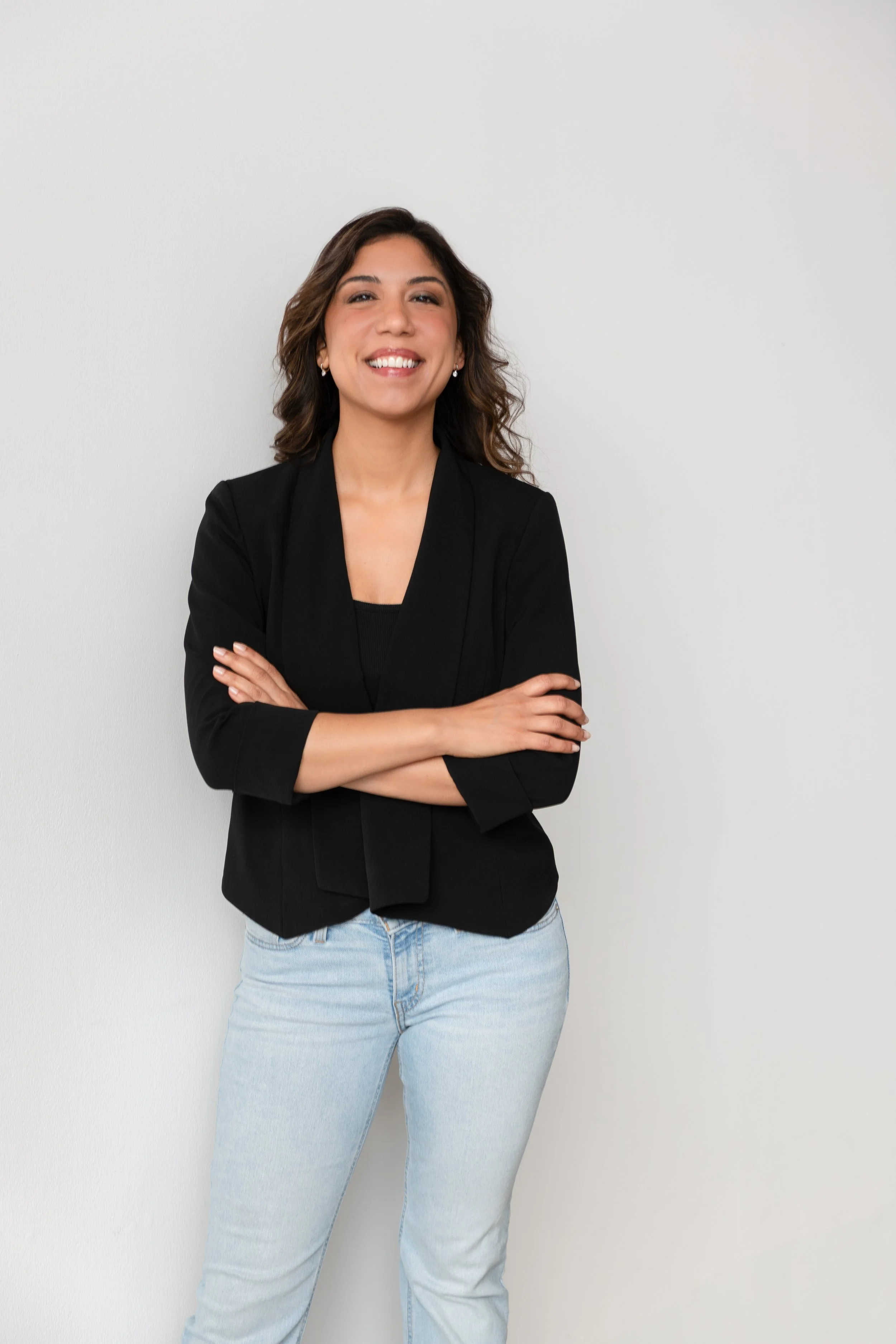 A woman with dark curly hair, wearing a black blazer and light blue jeans, standing with arms crossed and smiling, against a plain white wall.