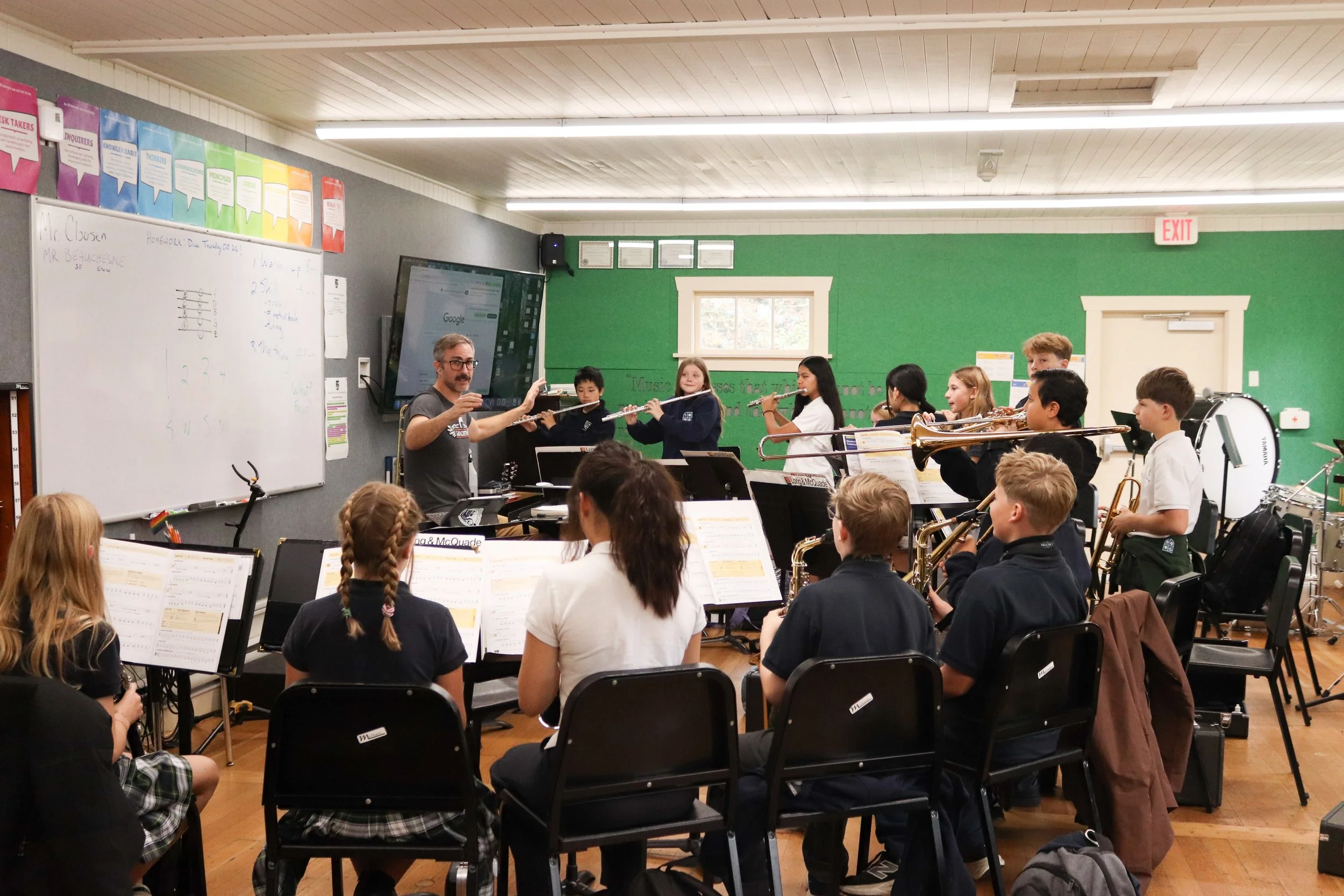 Music teacher leading a student band rehearsal with flutes, saxophones, trombones, and drums in a classroom