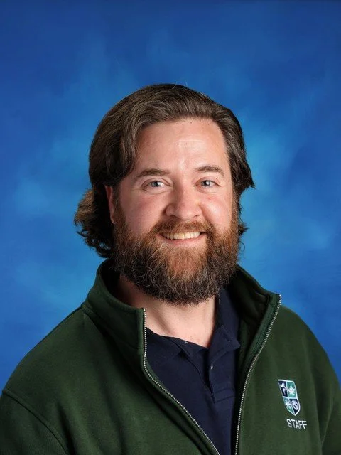 Portrait of a smiling bearded male staff member in a green jacket against a blue background