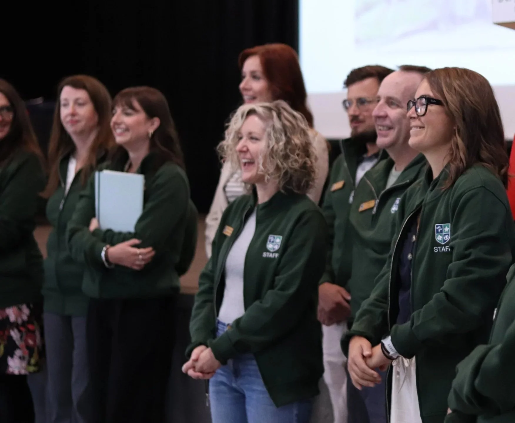 Group of smiling staff members in green jackets standing together at an indoor event