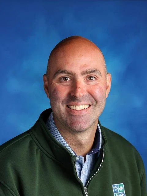 Smiling man wearing a green jacket over a blue checkered shirt against a blue studio background