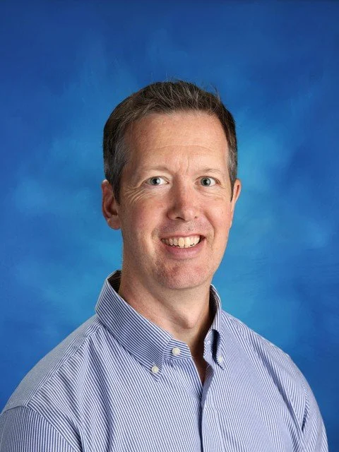 Professional headshot of a smiling man in a blue-and-white striped button-down shirt against a blue studio background