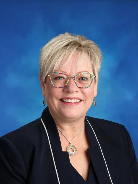 Professional headshot of a smiling woman with short blonde hair and colorful glasses wearing a black blazer against a blue background