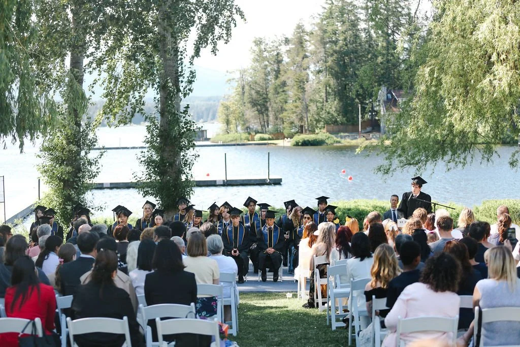 Outdoor graduation ceremony by a lake with graduates in caps and gowns and seated guests.