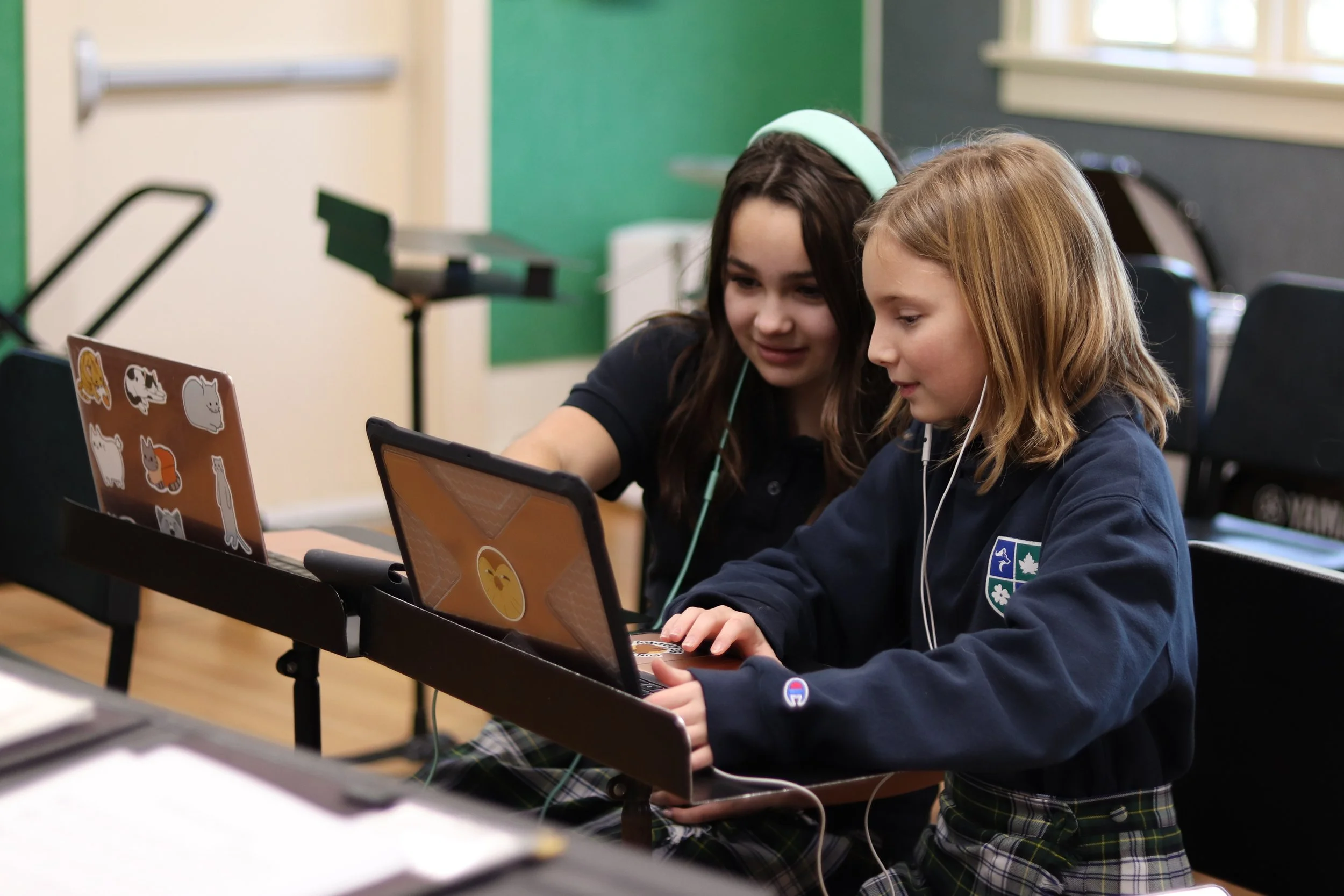 Two schoolgirls using laptops and a tablet together in a classroom learning music production.