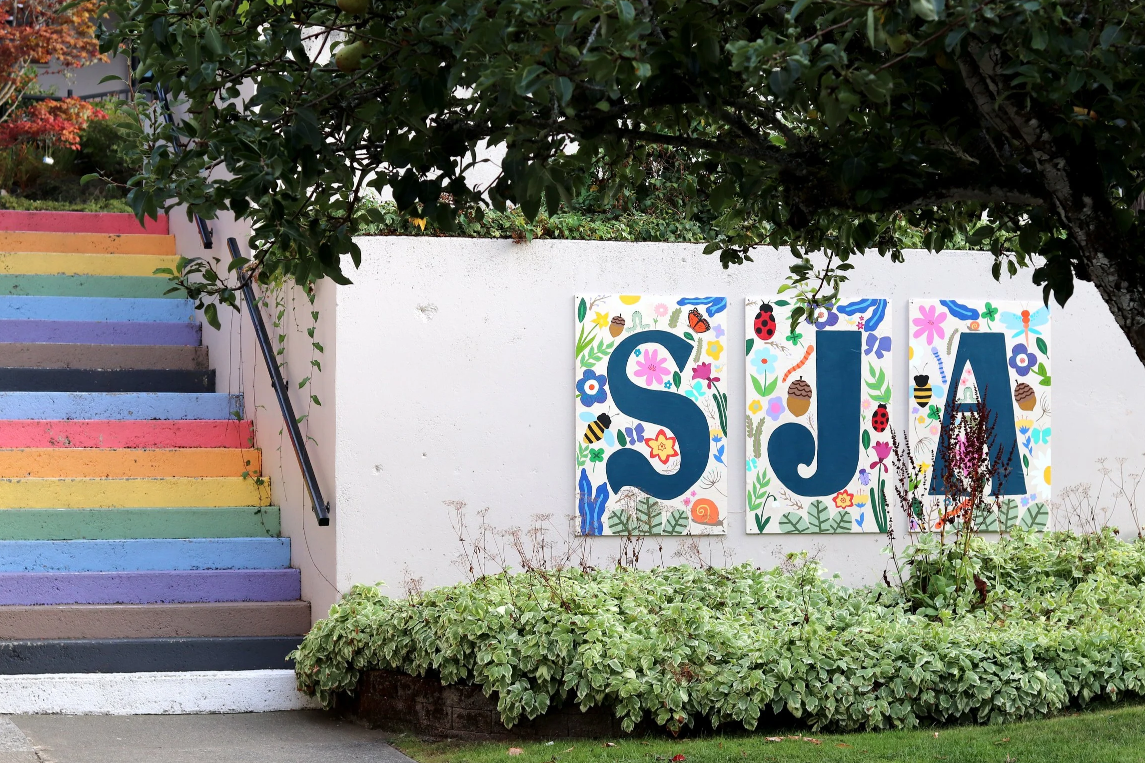 Colorful rainbow-painted steps and garden wall decorated with insect-and-flower artwork beneath a fruit tree