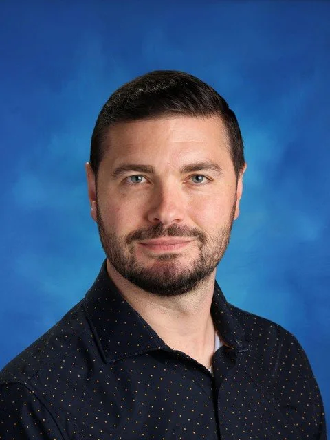 Professional headshot of a man in a dark patterned shirt against a blue studio background