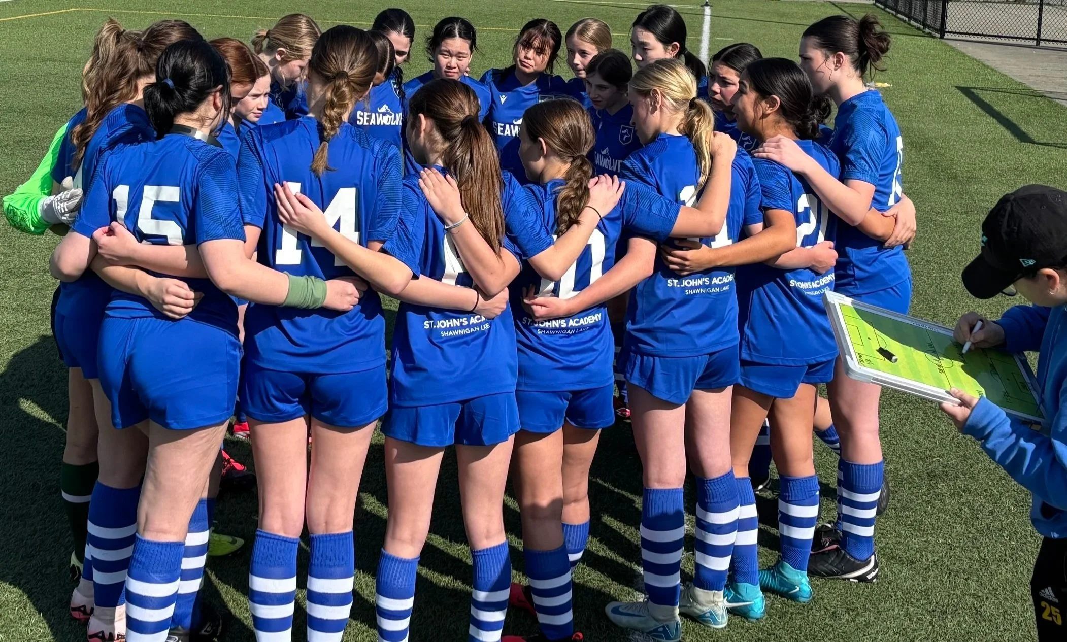 Girls' soccer team in blue uniforms huddles on a turf field as a coach reviews a strategy board.