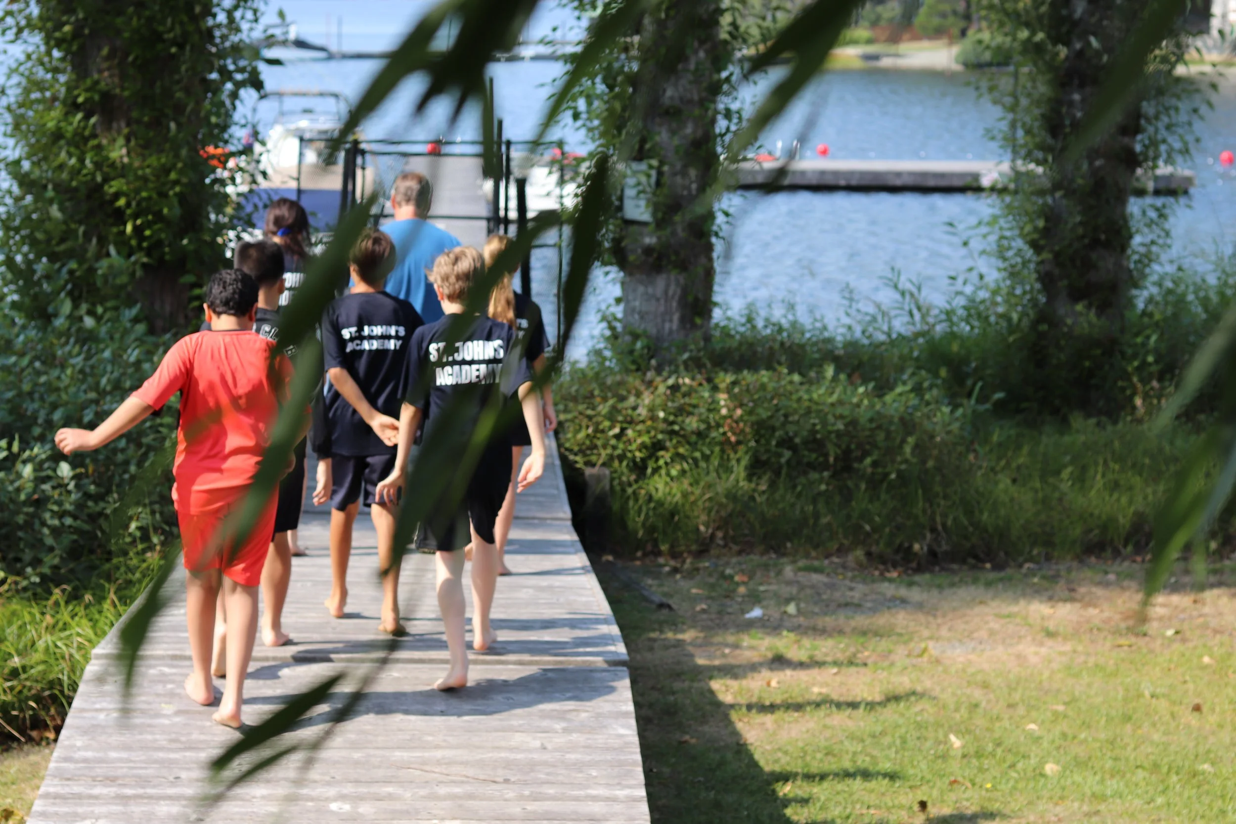 Children in St. John's Academy shirts walk barefoot along a wooded dock beside a lake.