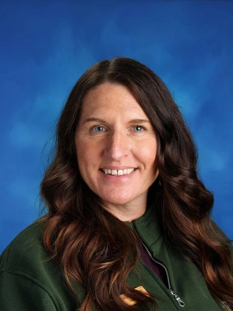 Professional headshot of a smiling woman with long wavy brown hair wearing a dark green jacket against a blue background.