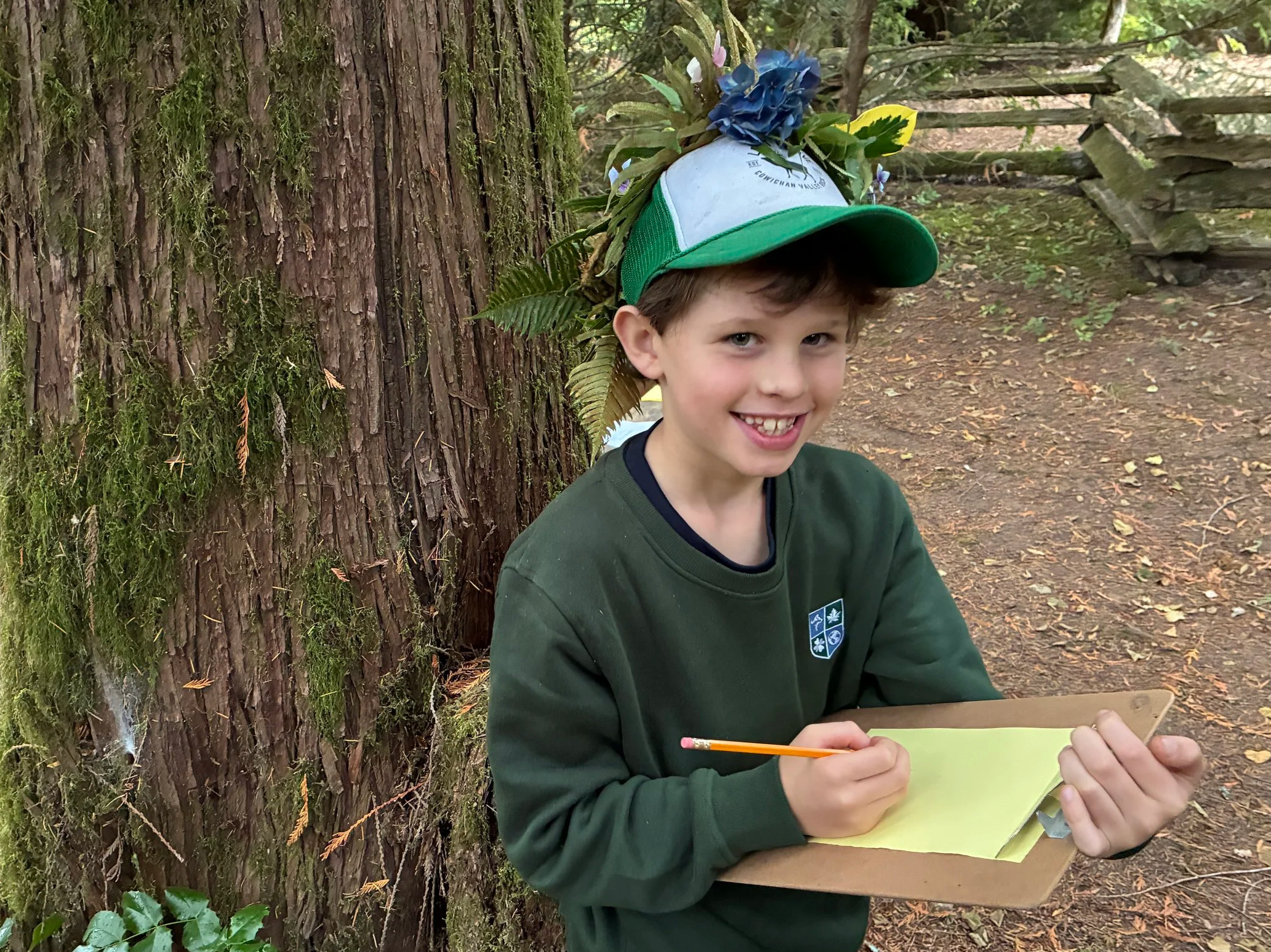 Smiling child wearing a flower-adorned green cap, writing on a clipboard beside a moss-covered tree in the woods
