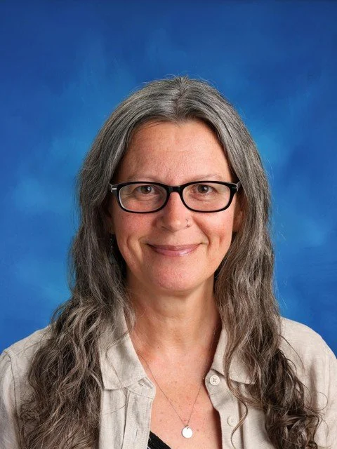 Professional headshot of a smiling woman with long gray hair and black glasses against a blue background
