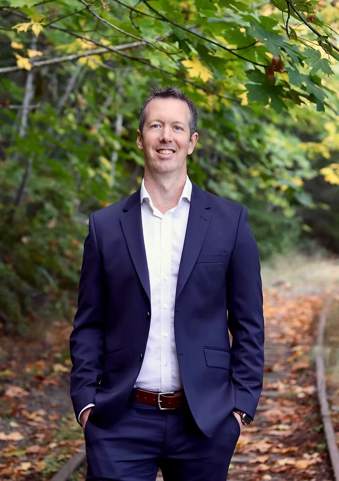 Smiling man in a navy suit and white shirt standing on a leafy woodland path