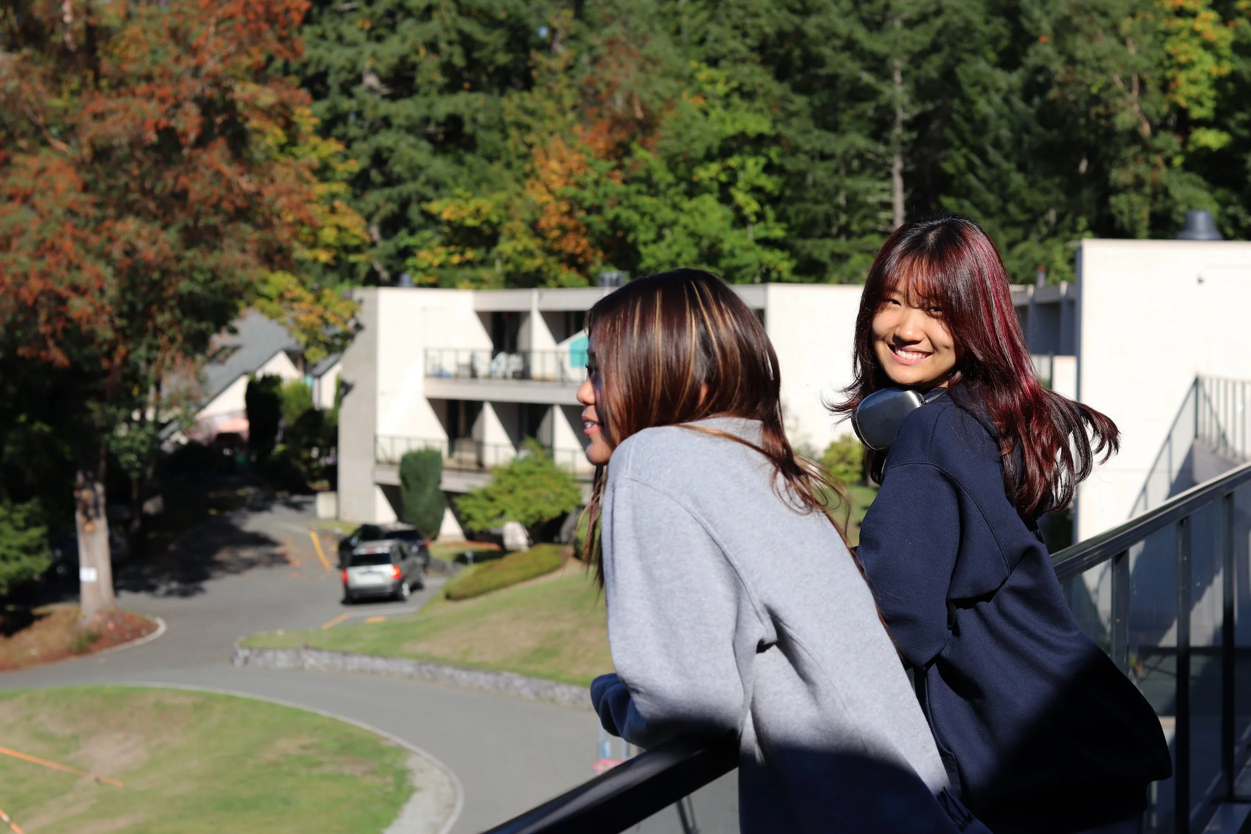 Two young women smiling on an outdoor balcony overlooking a tree-lined residential campus in autumn