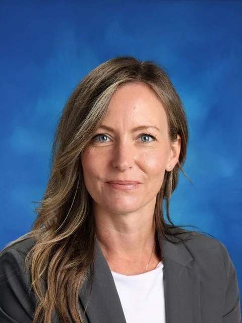 Professional headshot of a smiling woman with long brown hair wearing a gray blazer against a blue background