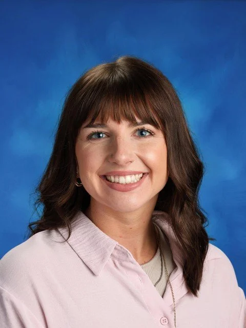 Professional headshot of a smiling woman with brown hair and bangs wearing a pink collared shirt against a blue background