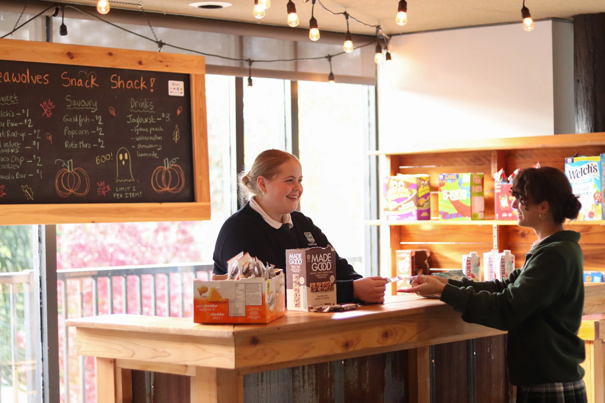 Two people exchange snacks at a cozy school snack shop counter with a chalkboard menu and shelves of packaged treats.