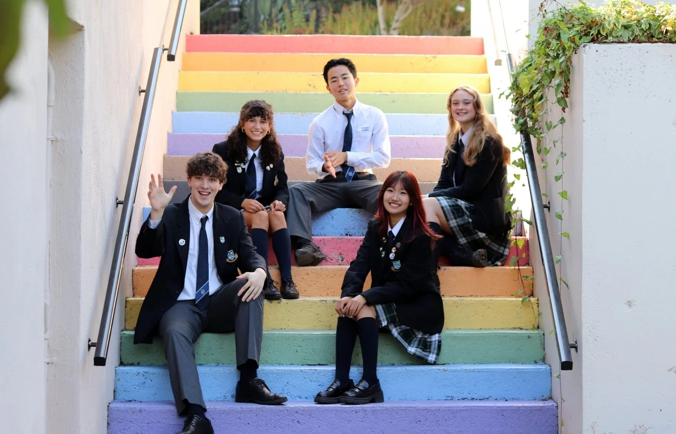 Five students in school uniforms pose on brightly painted rainbow-colored stairs.