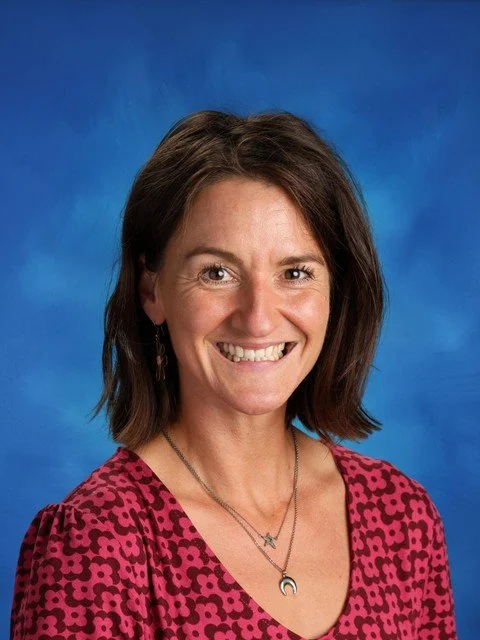 Smiling woman with shoulder-length brown hair wearing a pink patterned blouse and layered necklaces against a blue background