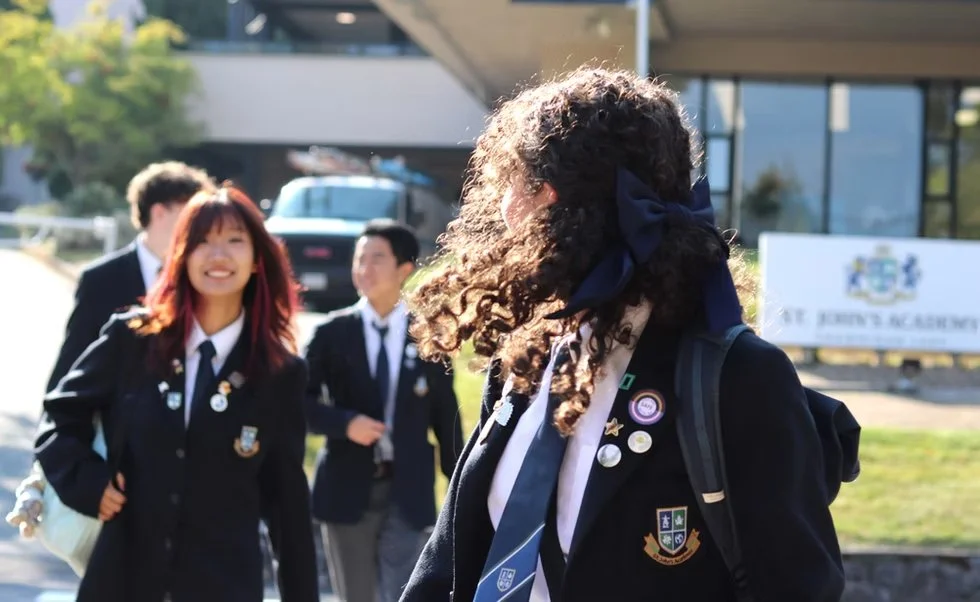 Uniformed students walking outside a school, with a curly-haired student wearing a large navy bow in the foreground.