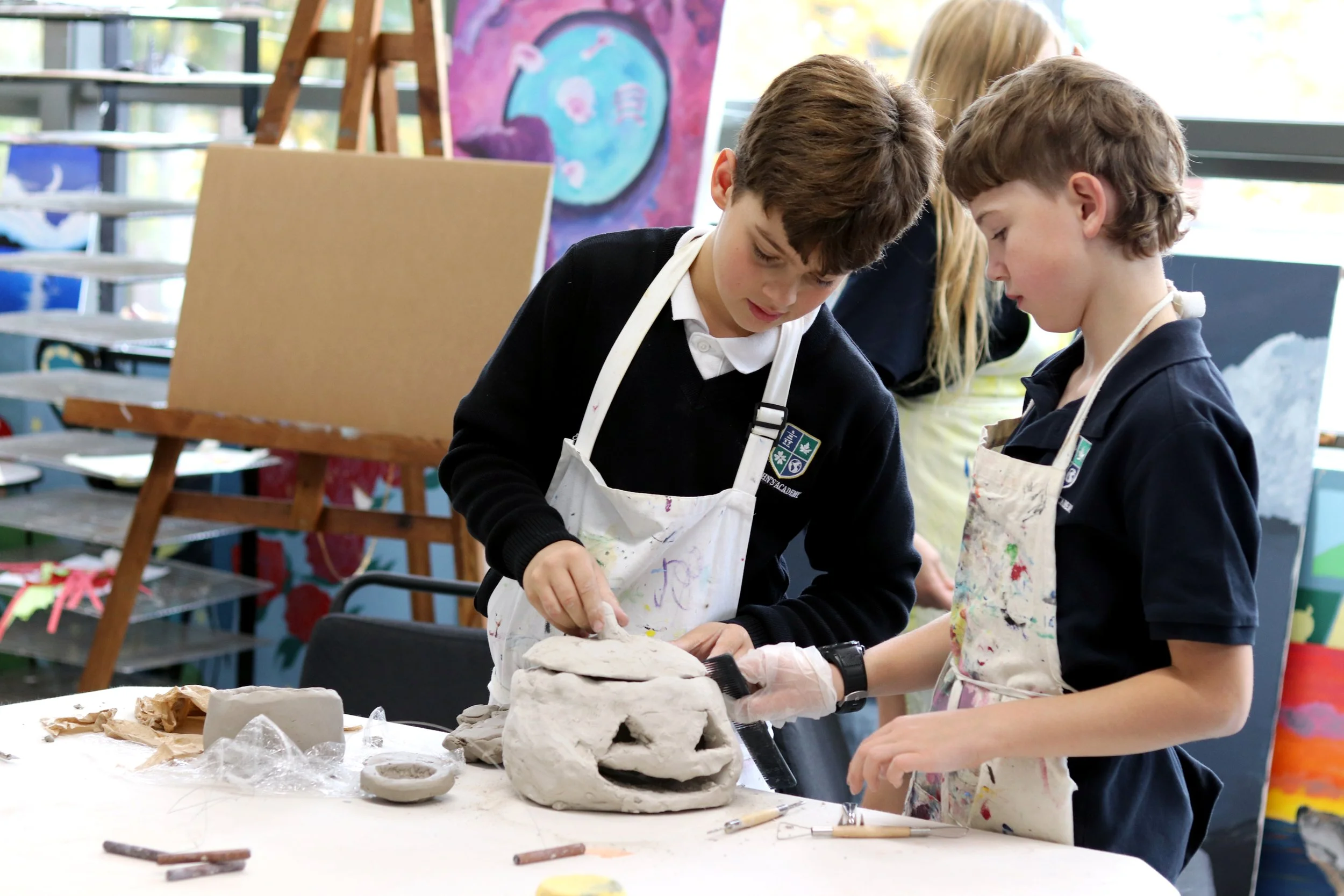 Students in art class wearing paint-splattered aprons and sculpting a clay project at a worktable.