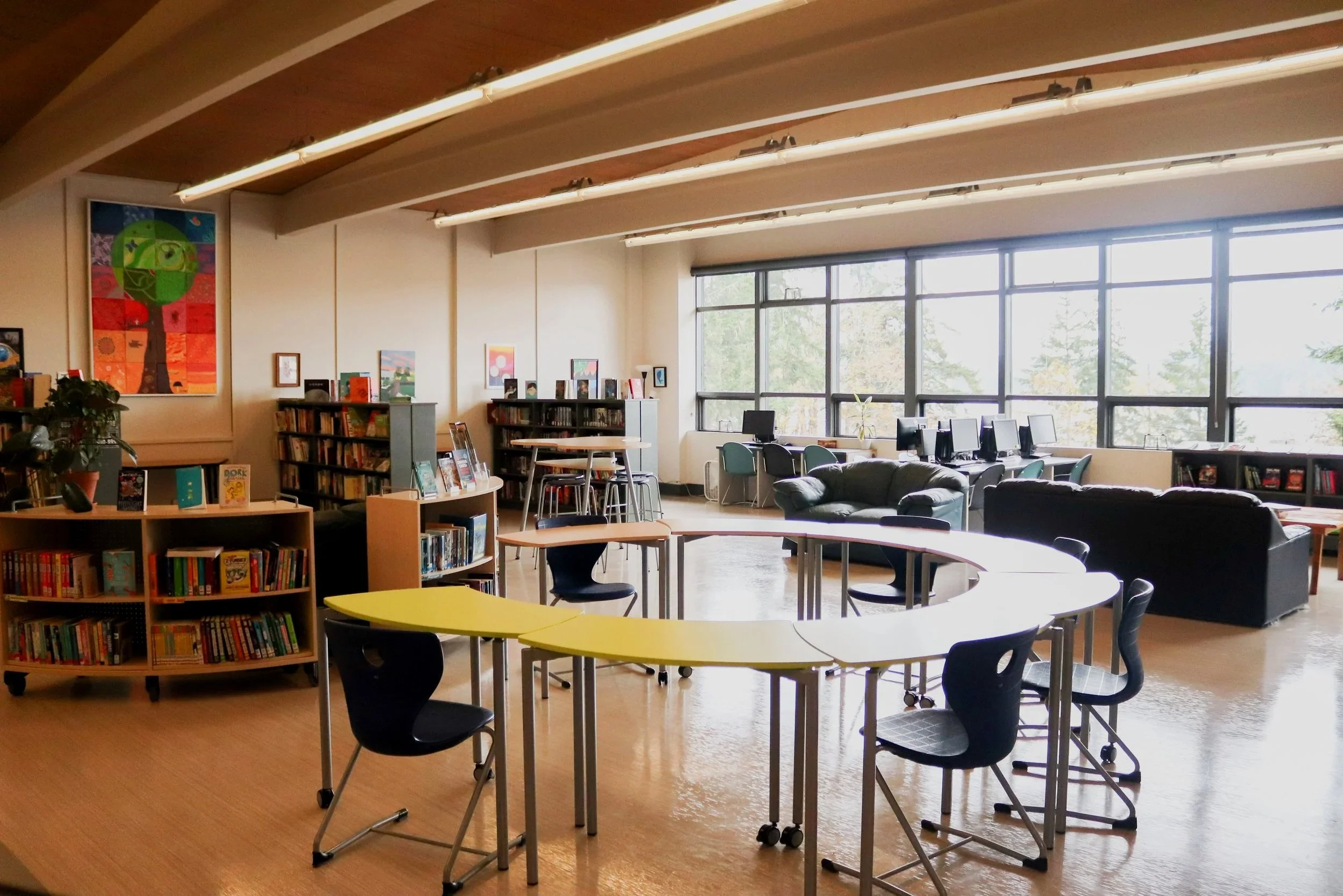 Bright school library with bookshelves, colorful tables and chairs, computers, couches, large windows, and wall artwork
