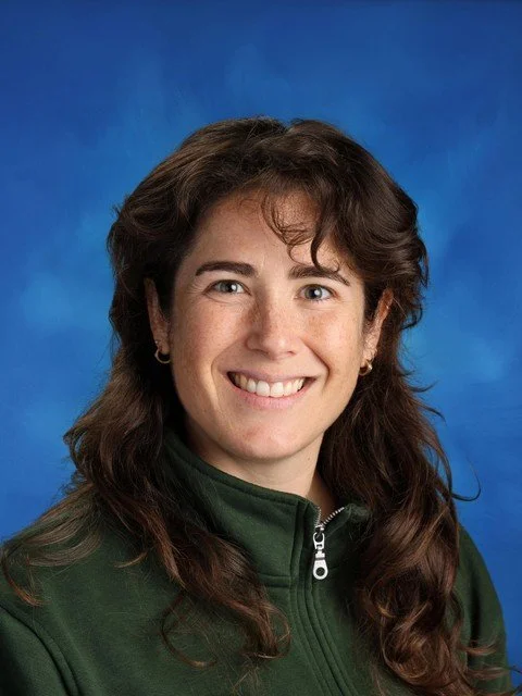 Smiling woman with long brown hair wearing a dark green zip-up jacket against a blue studio background