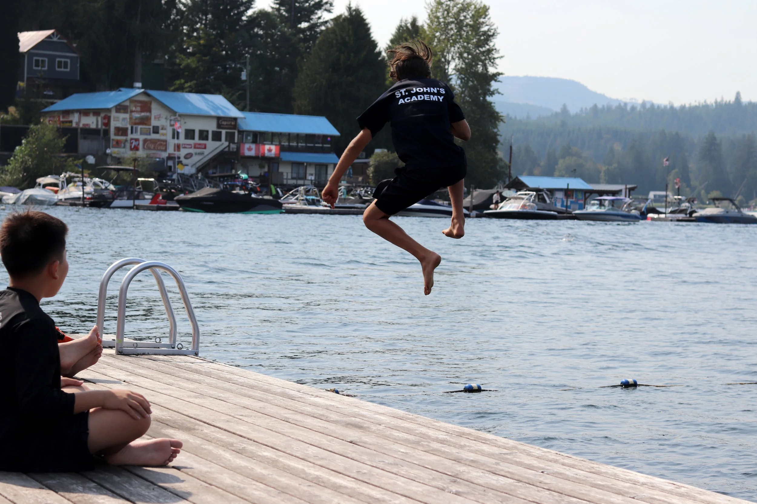 Child in a St. John's Academy shirt jumping from a dock into a lake, with boats, forested mountains, and a lakeside marina in the background