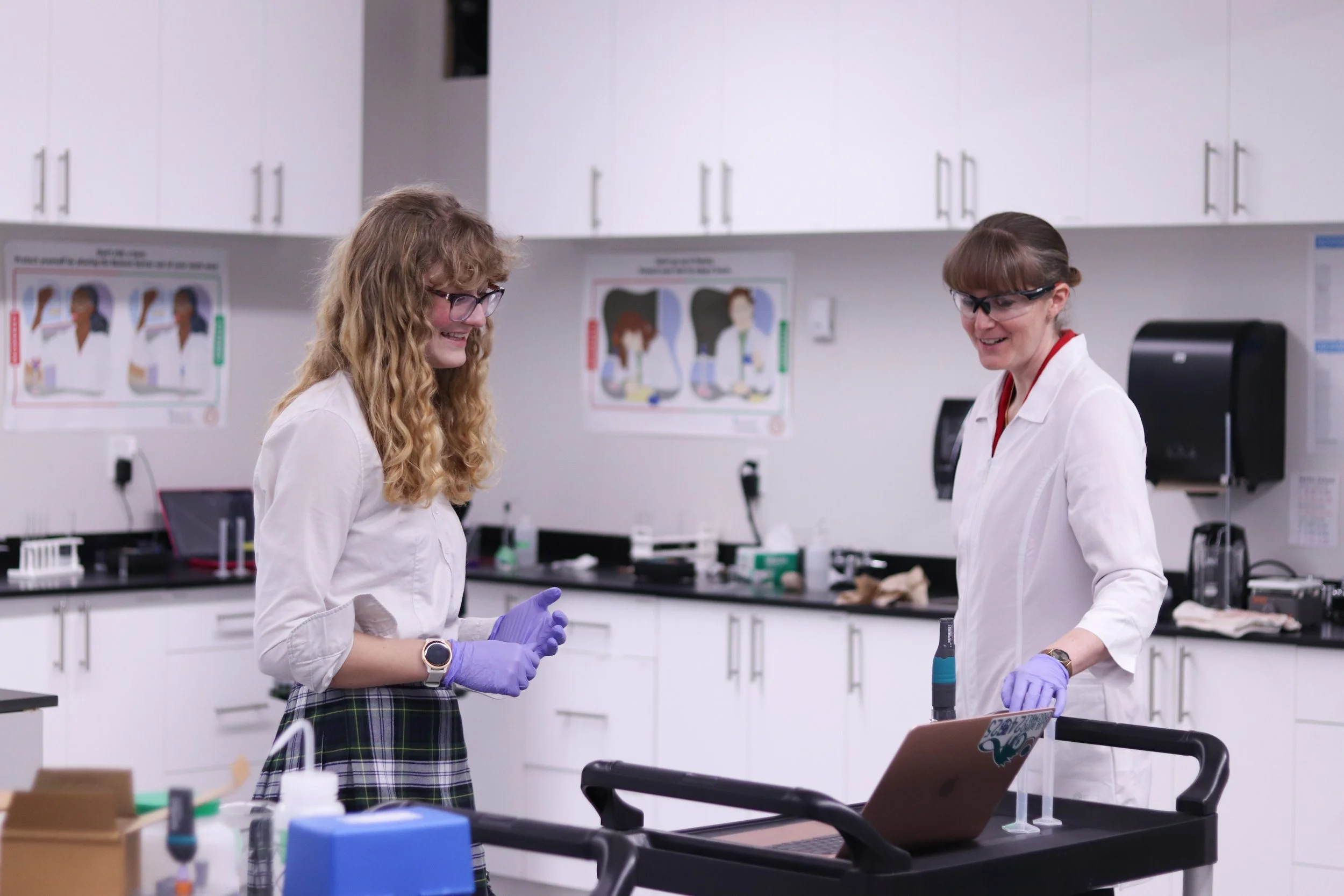 Two female scientists in lab coats and purple gloves working together in a modern laboratory.