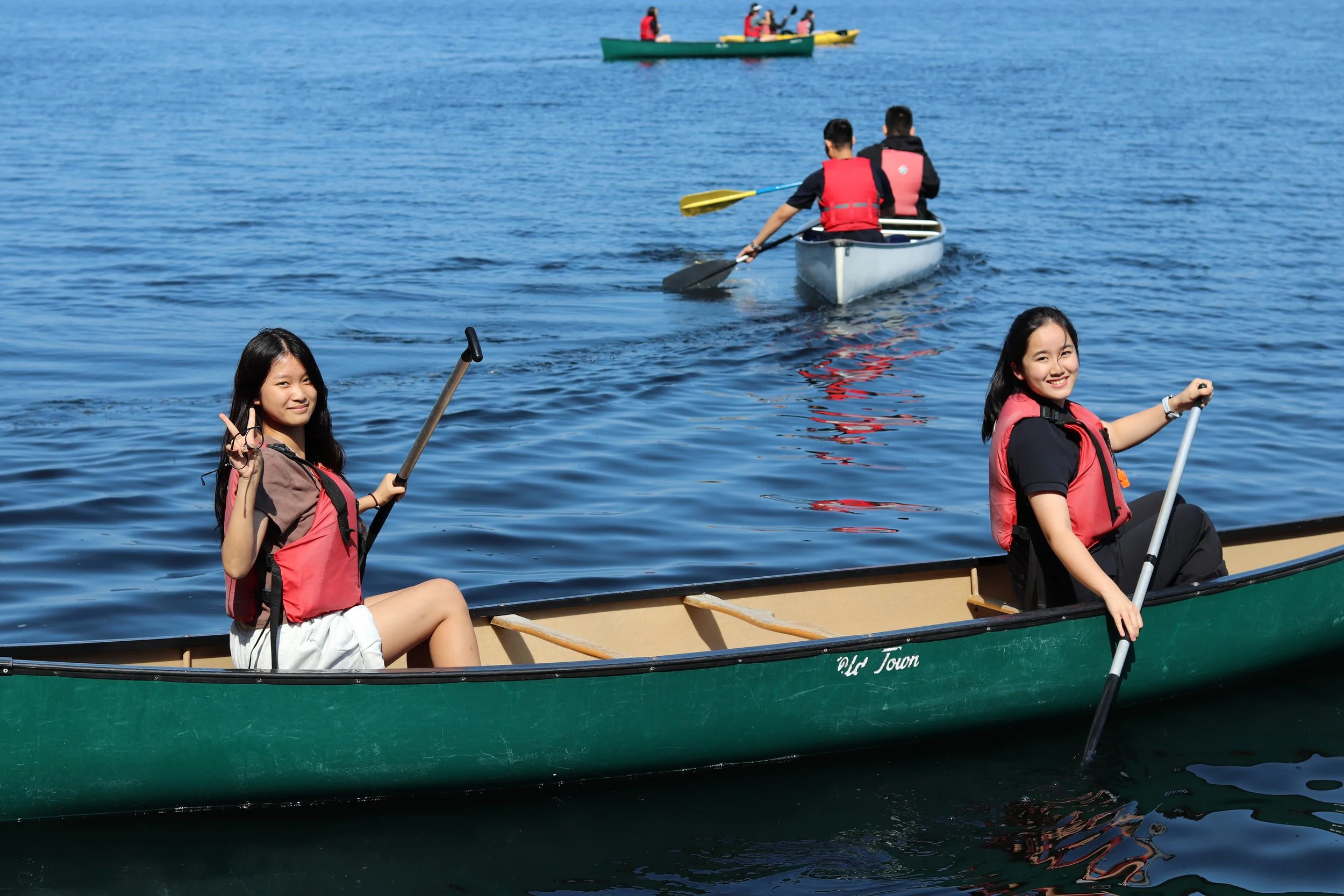 People wearing life jackets canoeing on a blue lake, with paddlers in a canoe in the foreground and background.
