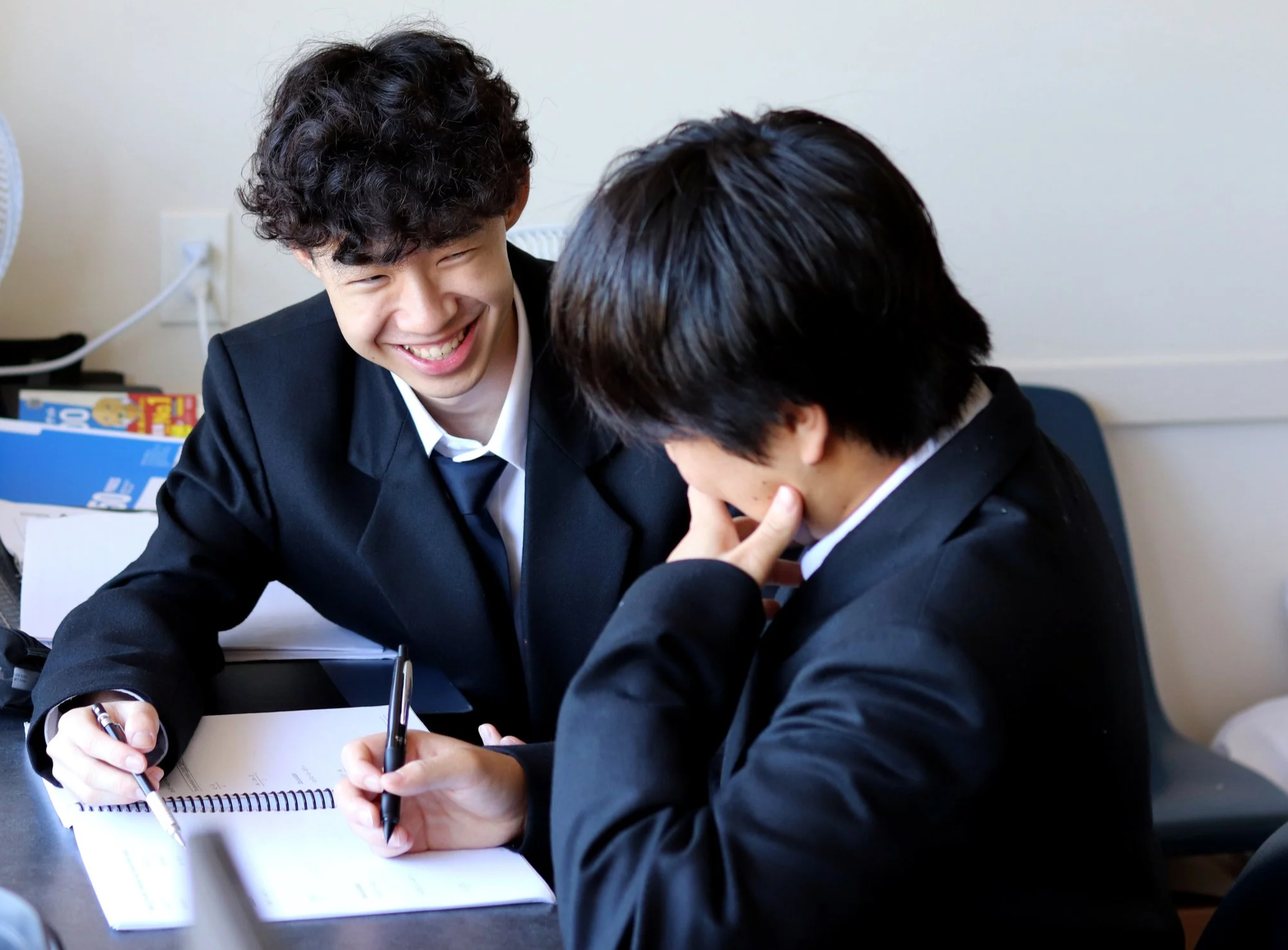 Two students in black suits laughing and writing together in a notebook at a desk