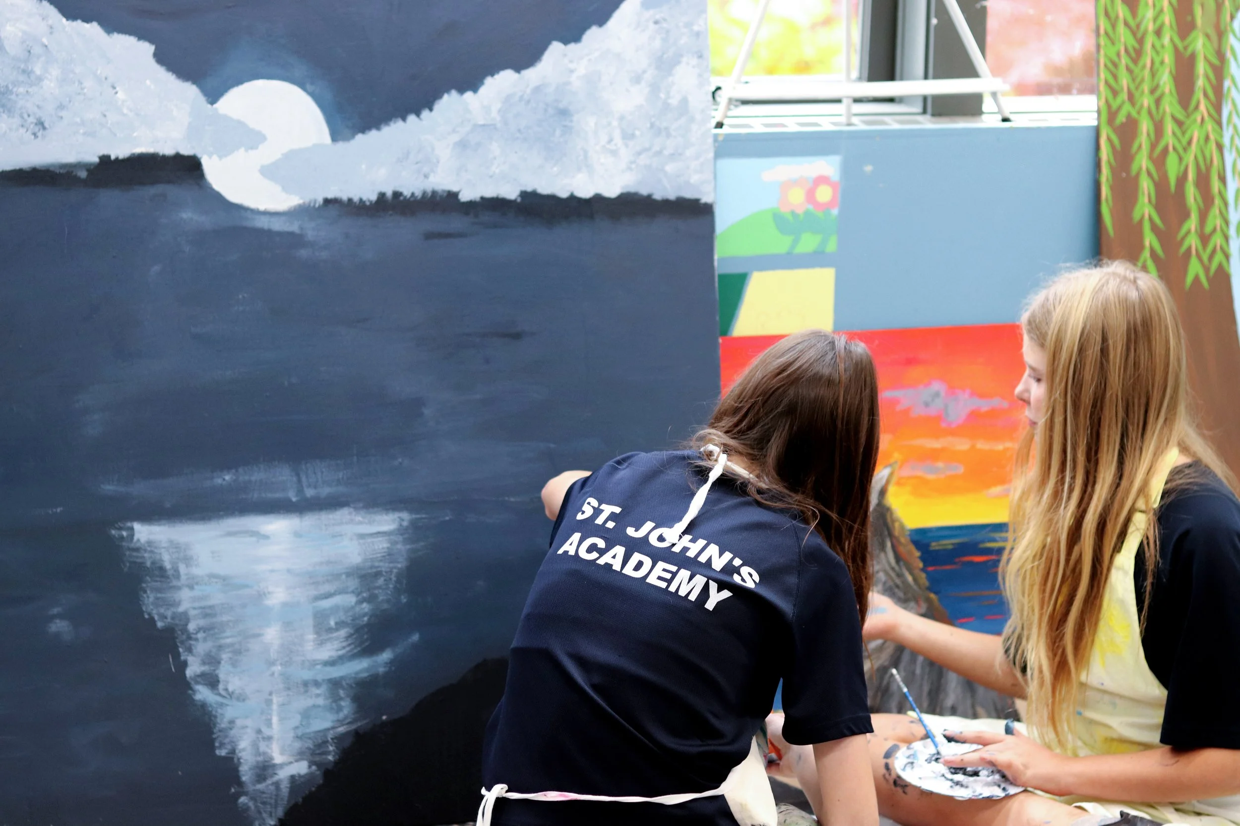 Students paint a large mural beside a colorful outdoor artwork, with one wearing a St. John’s Academy shirt.