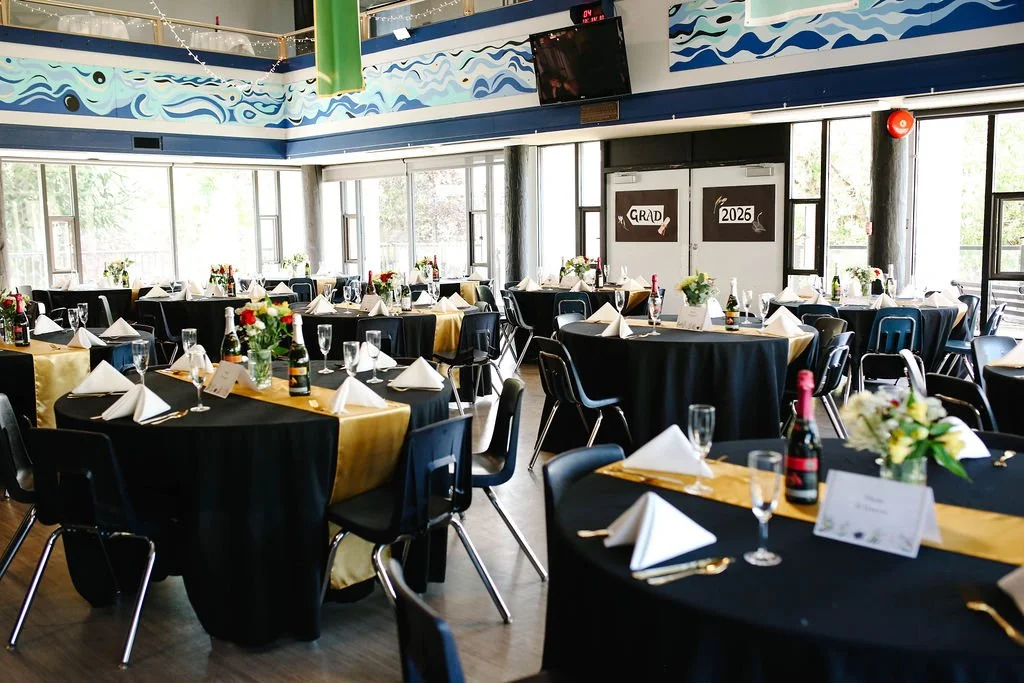 Banquet hall decorated for a graduation celebration with tablecloth-covered tables, folded napkins, flowers, champagne, and place settings.