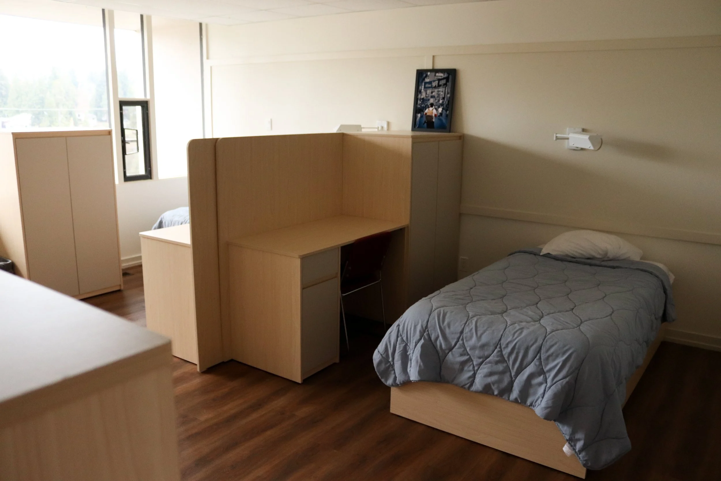 Modern bedroom with a gray quilted bed, light wood partition and cabinets, large windows, and hardwood flooring