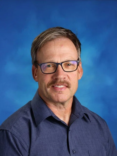 Professional headshot of a man wearing glasses and a dark blue collared shirt against a blue studio background
