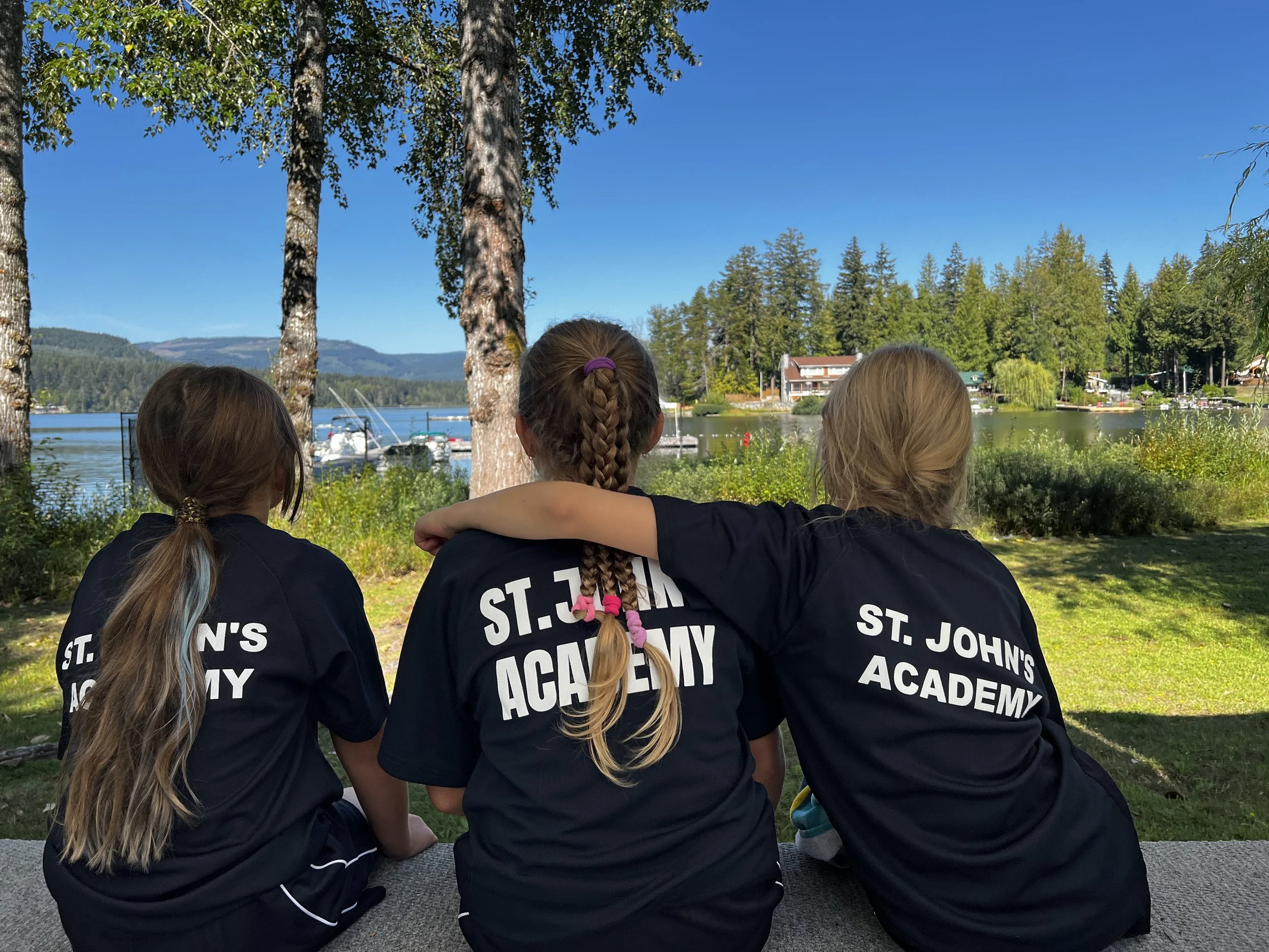 Three girls wearing St. John's Academy shirts sit beside a lake with boats, trees, and mountains under a clear blue sky.