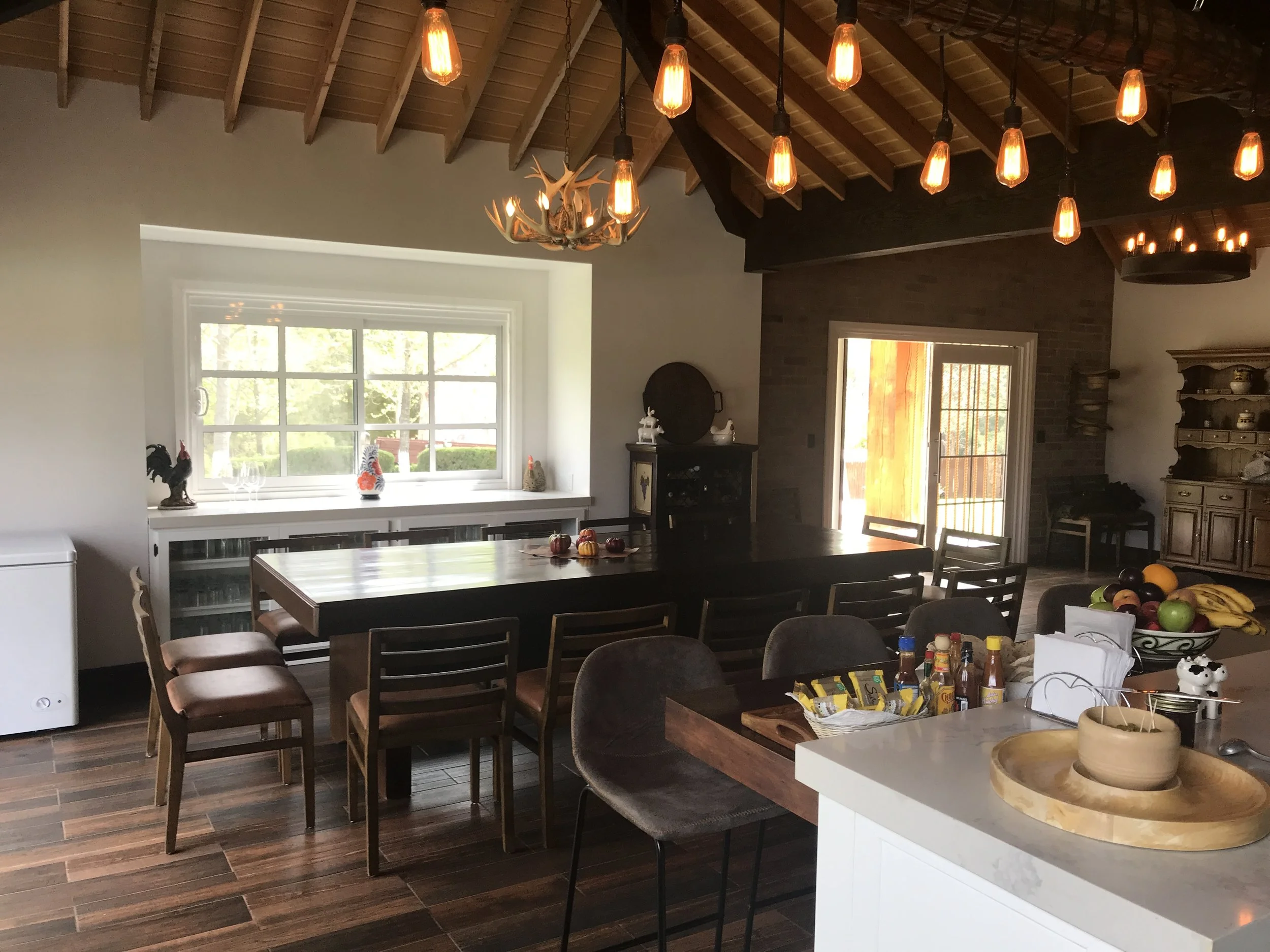 Open-plan kitchen, dining and living area with wooden ceiling beams, pendant lights, and views through the window.